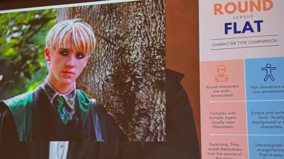 Person with blonde hair and dark eyeliner stands outdoors by a tree. A poster comparing round and flat character types is partially visible.