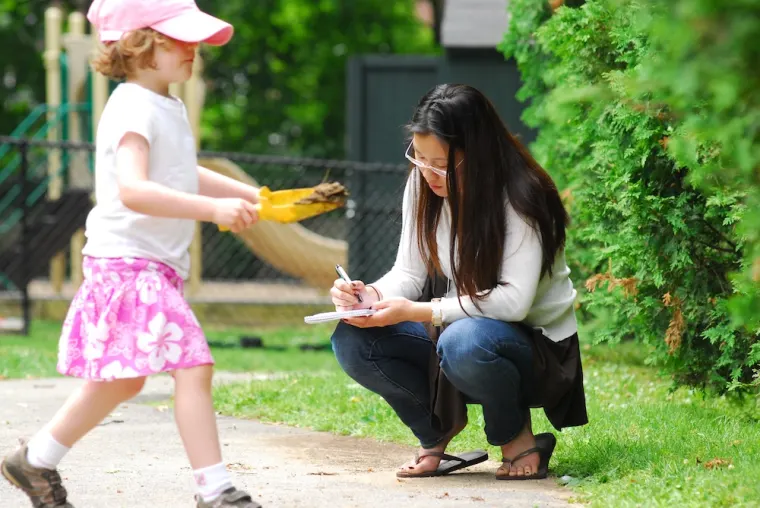 Adult crouching and writing on notepad while child in pink runs by.