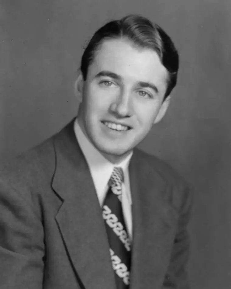Person in suit with patterned tie smiling against plain background.
