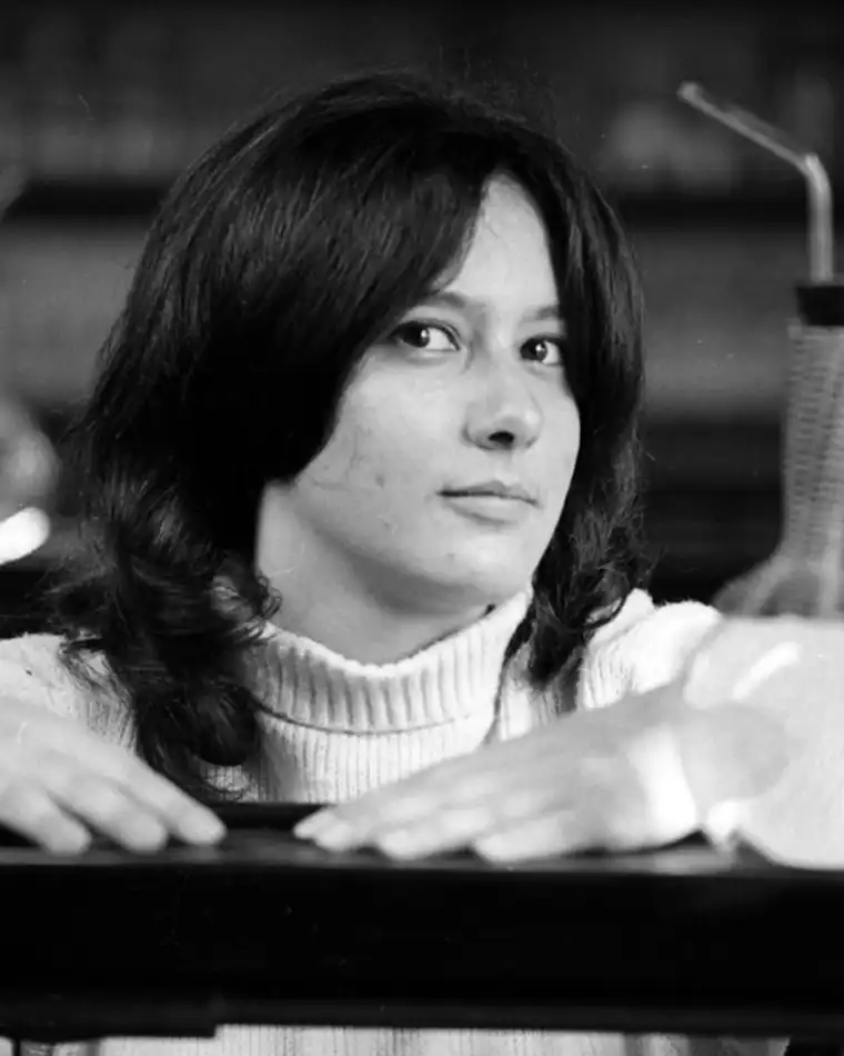 Individual with long hair in a turtleneck, resting arms on a surface, with a thoughtful expression. Black and white photo.