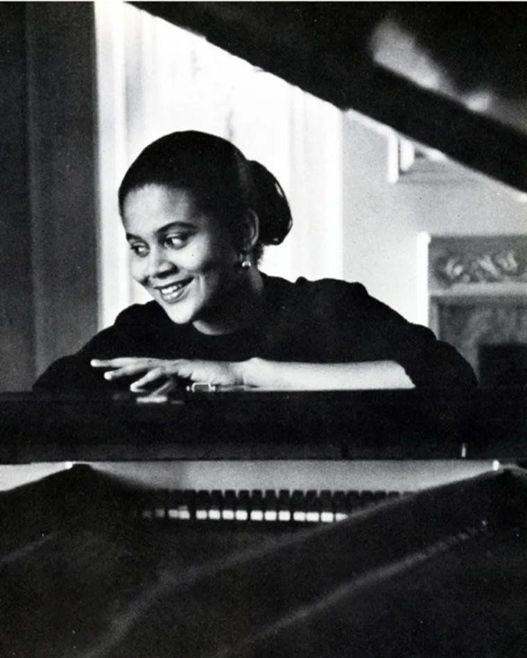 Person smiling and resting hands on a piano, viewed between piano lid and keyboard.
