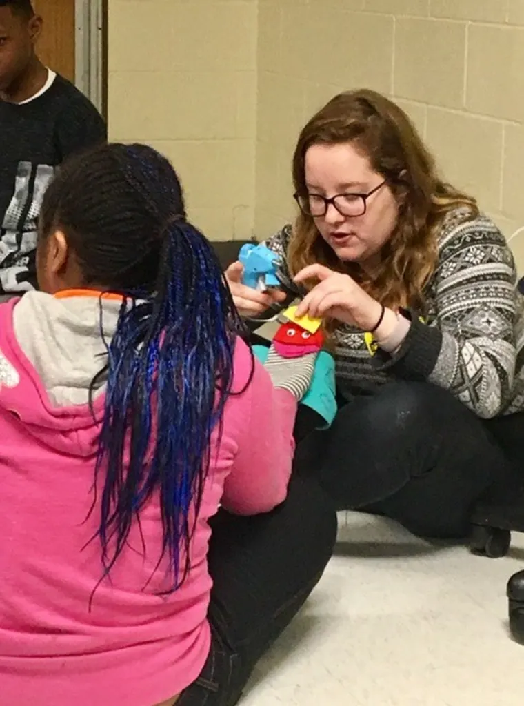 Adult showing a child how to sew a blue fabric piece, indoor setting.