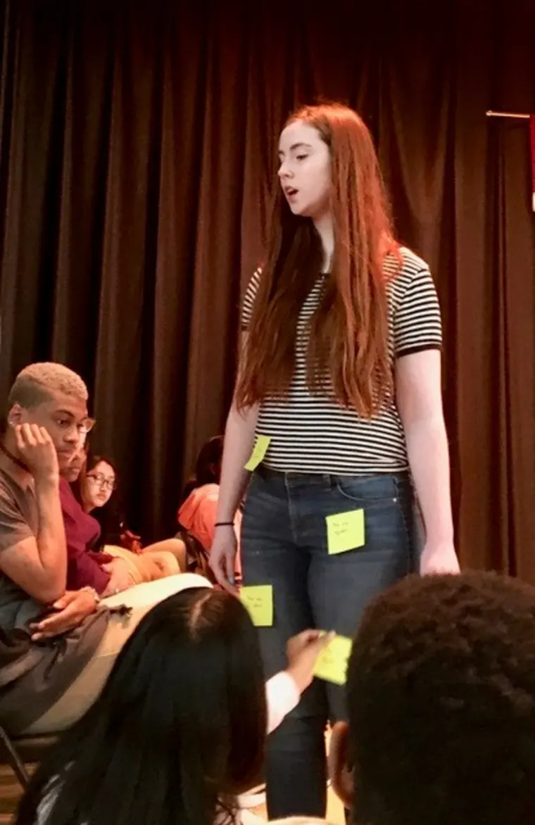 Person speaking on stage with audience members looking on.