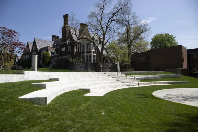 A Tudor-style house with a large brick chimney behind an open grassy area with modern white tiered seating steps on a sunny day.