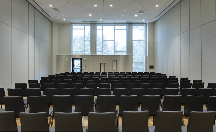 A modern conference room with rows of black chairs facing a podium at the front, large windows providing natural light, and a neutral color scheme.