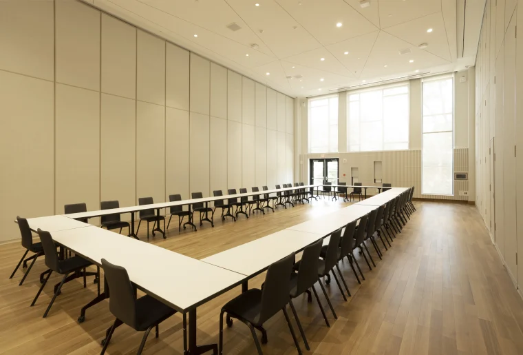 A spacious and modern meeting room with a long arrangement of tables set up in a U-shape, surrounded by numerous chairs, under ceiling lights. Large windows provide natural light, and the room features a minimalist design with light-colored walls and wooden flooring.
