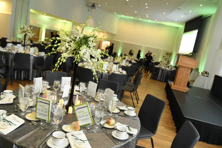 Elegantly set banquet hall with multiple round tables arranged with dinnerware, glassware, and floral centerpieces, with a stage and podium in the background.