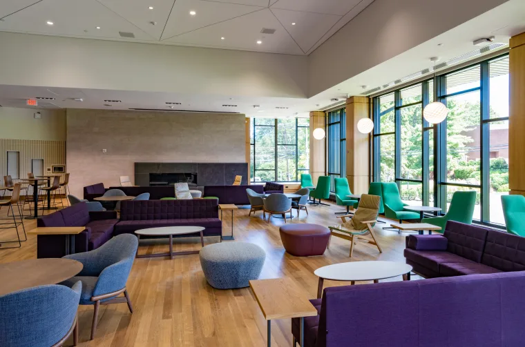 Modern lounge area with a variety of seating options including couches and chairs in shades of green and purple, large windows providing natural light, and a fireplace on one wall.