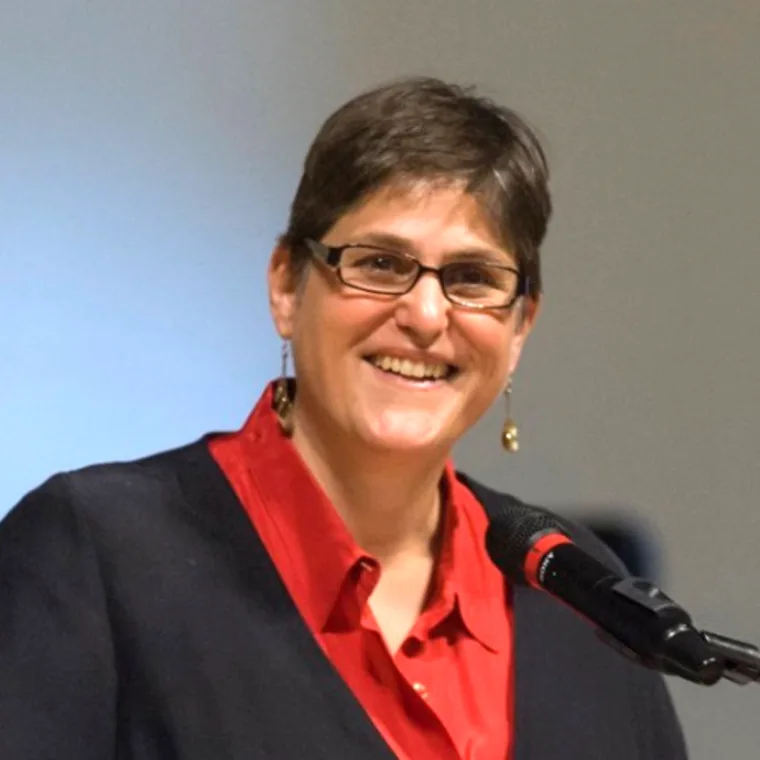 Person with short hair and glasses smiles while speaking into a microphone, wearing a red shirt and dark jacket.