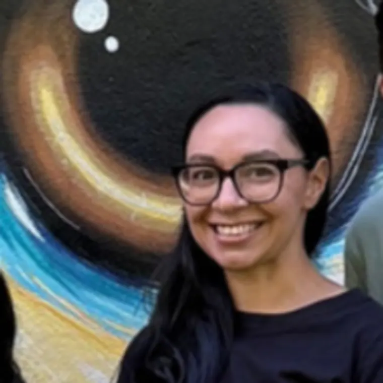 Person smiling in front of vibrant mural with large eye design, showing shades of orange, blue, and white.