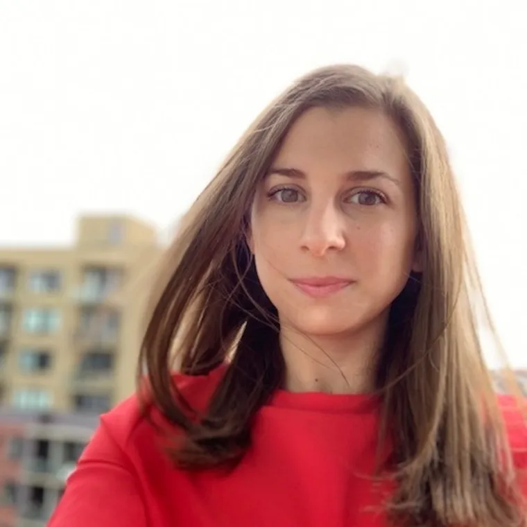 Person with long hair wearing a red shirt, standing outdoors with blurred buildings in the background.