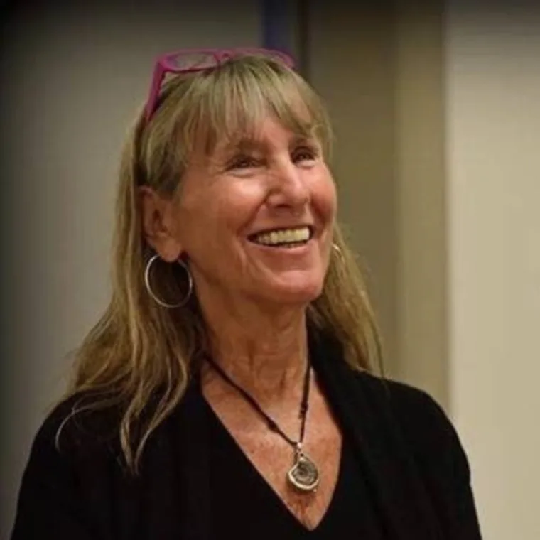 Person smiling, wearing hoop earrings and a pendant necklace, with glasses on top of their head.