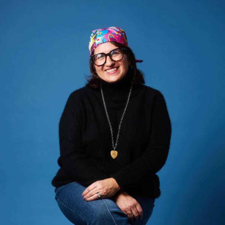 Person smiling, wearing glasses, a vibrant headscarf, black sweater, blue jeans, and a pendant necklace, against a solid blue background.