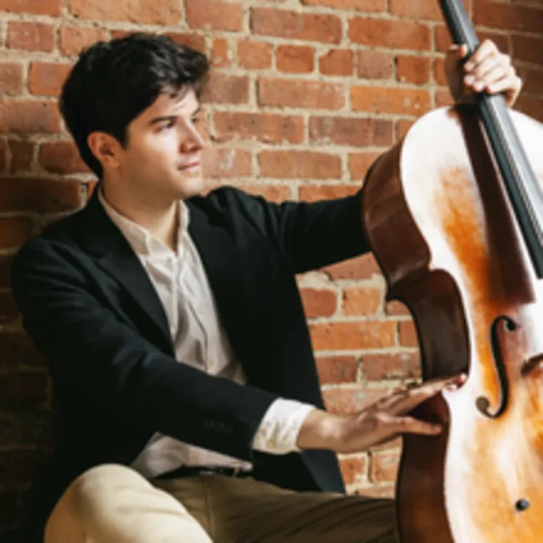 Person in a blazer playing a cello against a brick wall background.