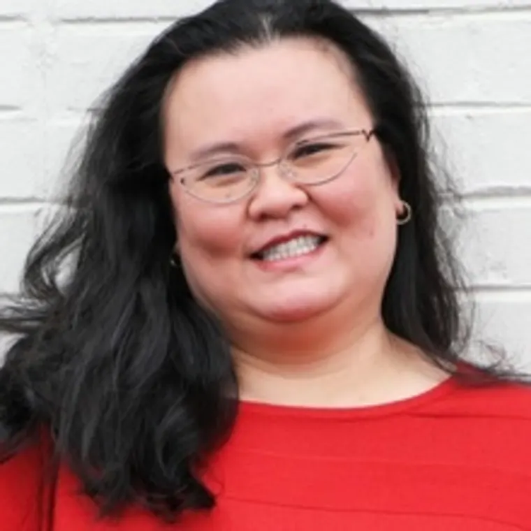 Person with long black hair and glasses smiling, wearing a red shirt in front of a white brick wall.