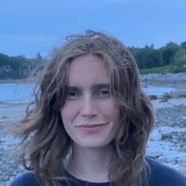 Person with long hair smiling at a beach, with trees and water in the background.