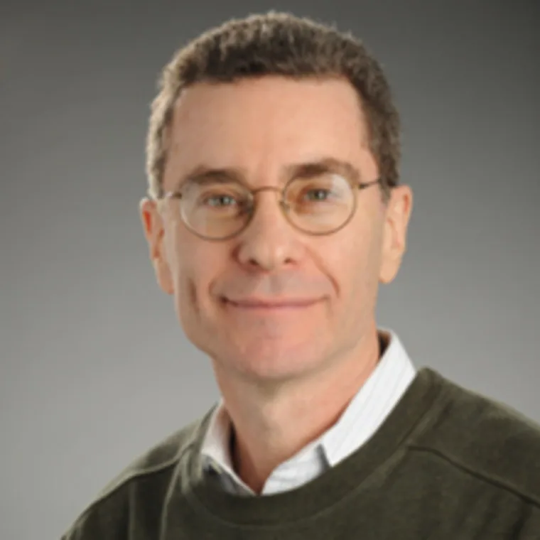 Person with short hair and glasses, wearing a dark sweater over a collared shirt, smiles at the camera against a gray background.