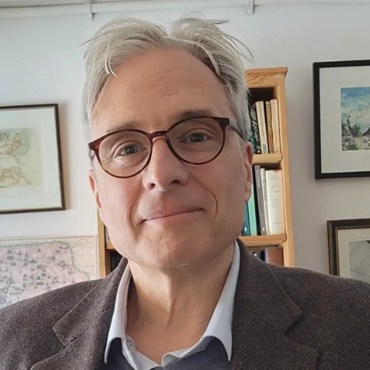 Person with glasses and grey hair, wearing a blazer. Standing in a room with framed pictures on the wall and books on a shelf.