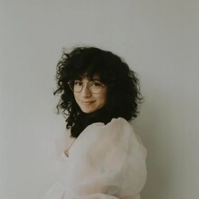 Person with curly hair and glasses, wearing a light, sheer top, smiles at the camera against a plain background.