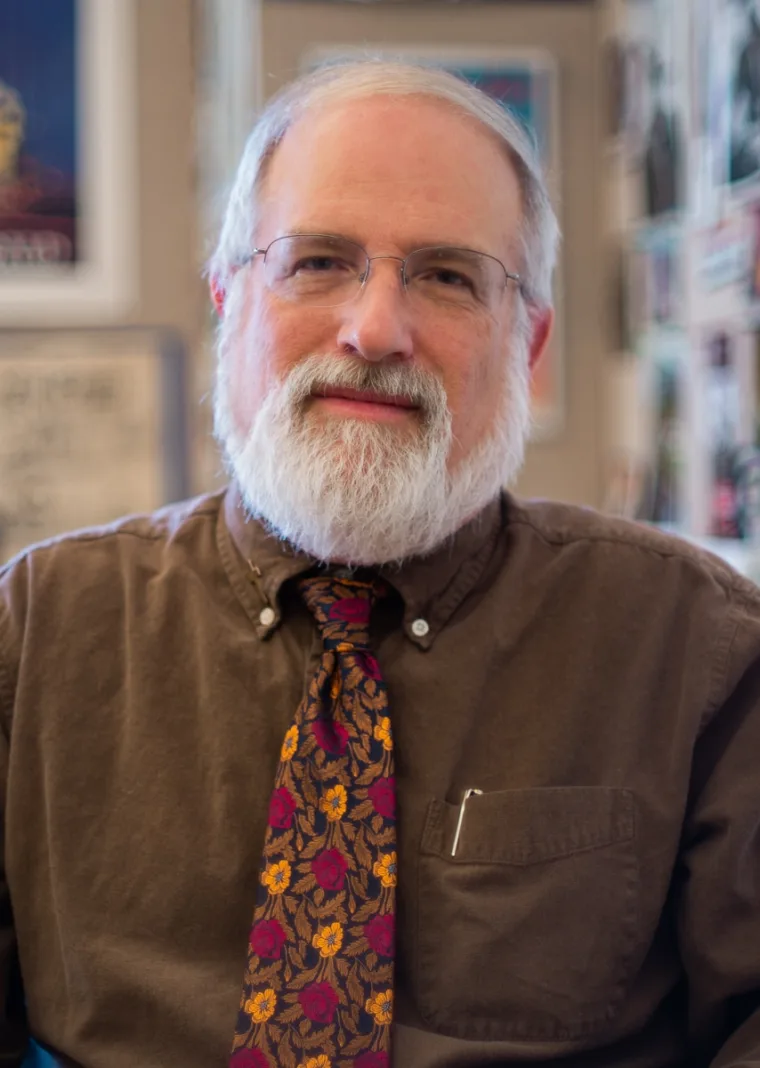 Person with white beard and glasses wearing a brown shirt and floral tie, sitting indoors with blurred decorations in the background.