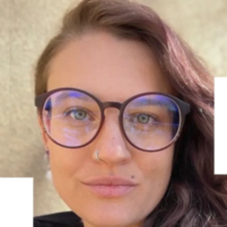 Person with long hair, wearing large round glasses, nose ring, and a mole above the lip, looking at the camera with a neutral expression.