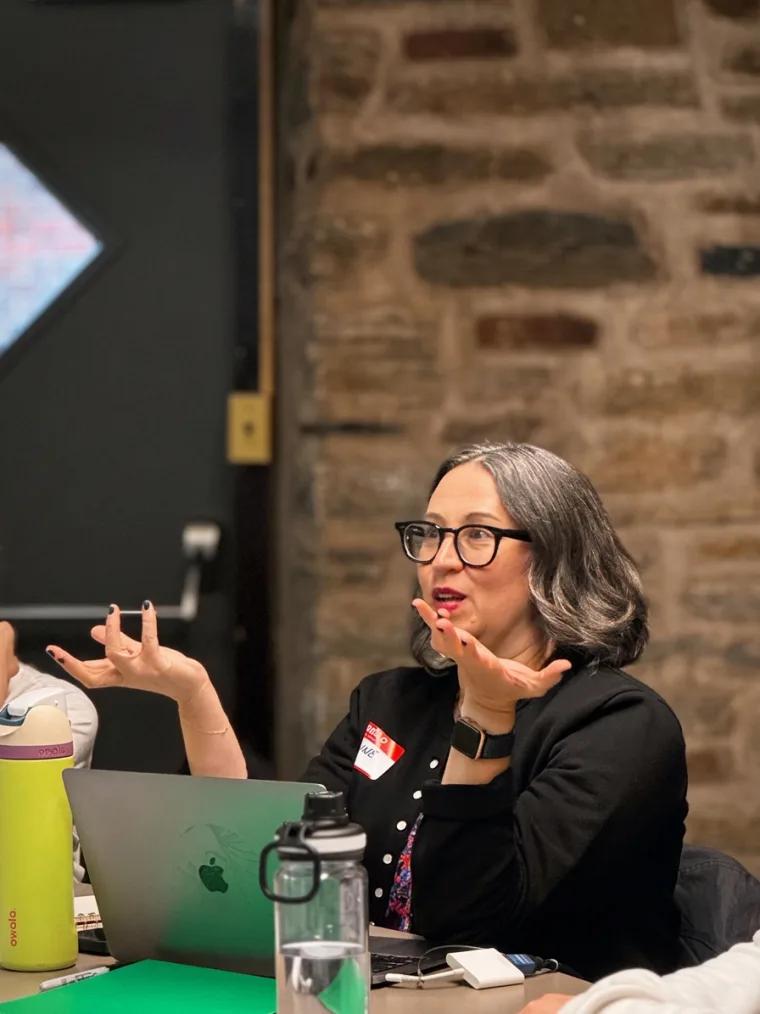 Person with glasses speaking animatedly, seated at a table with a laptop and water bottle. Brick wall and door in the background.