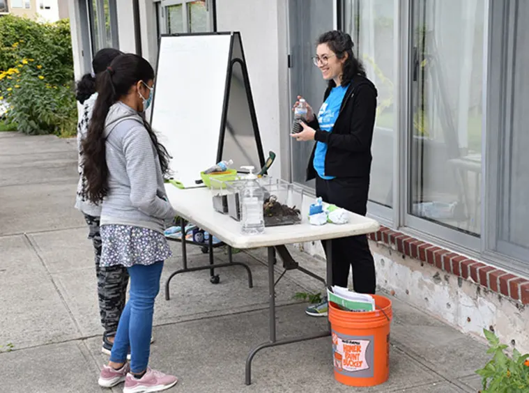 Person demonstrating a science experiment outdoors at a table, with two others observing. Supplies and a flip chart are on the table.