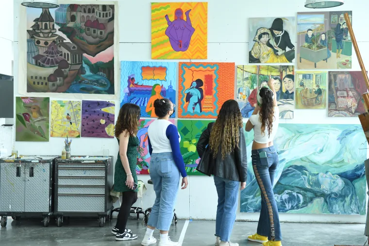 Four people observing colorful paintings on a studio wall, with an easel and art supplies nearby.