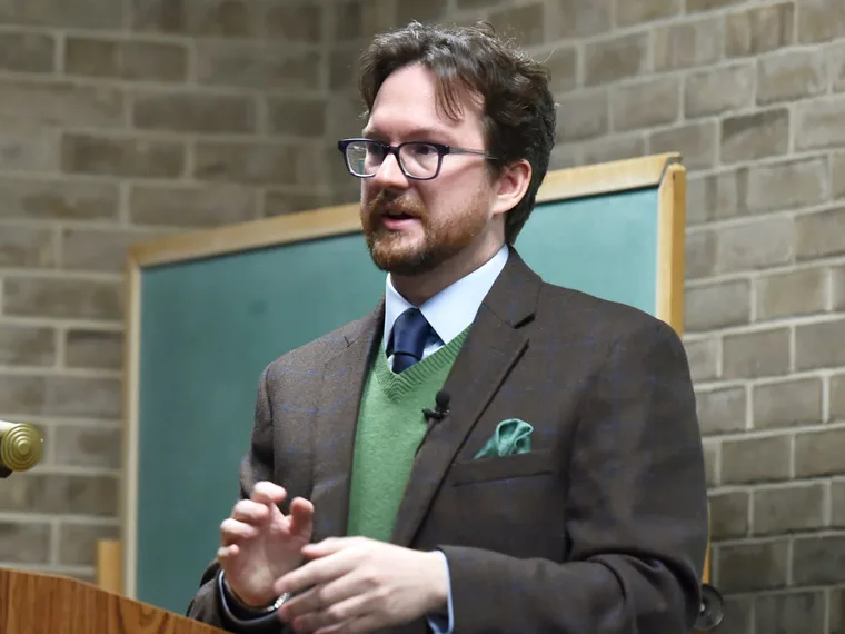 Person wearing glasses and a suit with a green sweater speaks at a lectern, with a chalkboard and brick wall in the background.