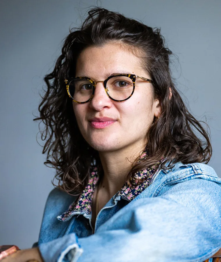 Person with curly hair, wearing tortoiseshell glasses and a denim jacket, looks at the camera against a plain background.