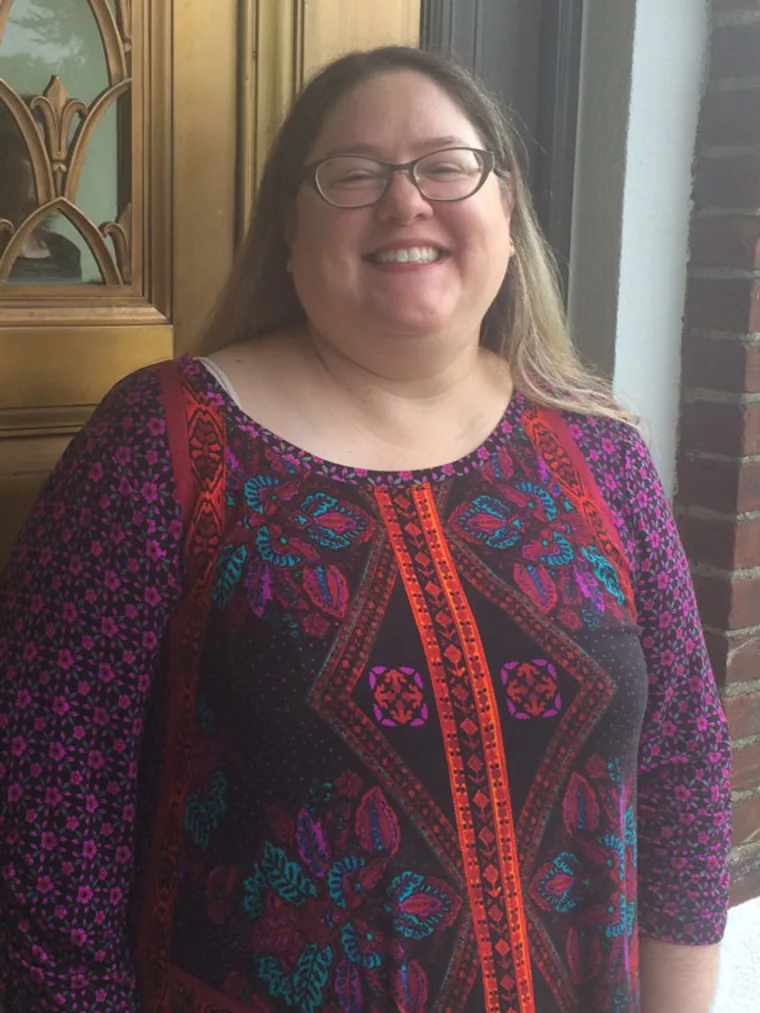 Person smiling in front of a wooden door, wearing glasses and a colorful patterned top.