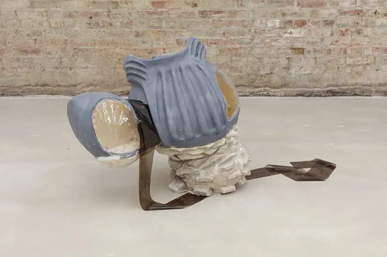Abstract sculpture with gray and beige elements, resembling an intricate, futuristic armor piece on a beige floor against a brick wall.