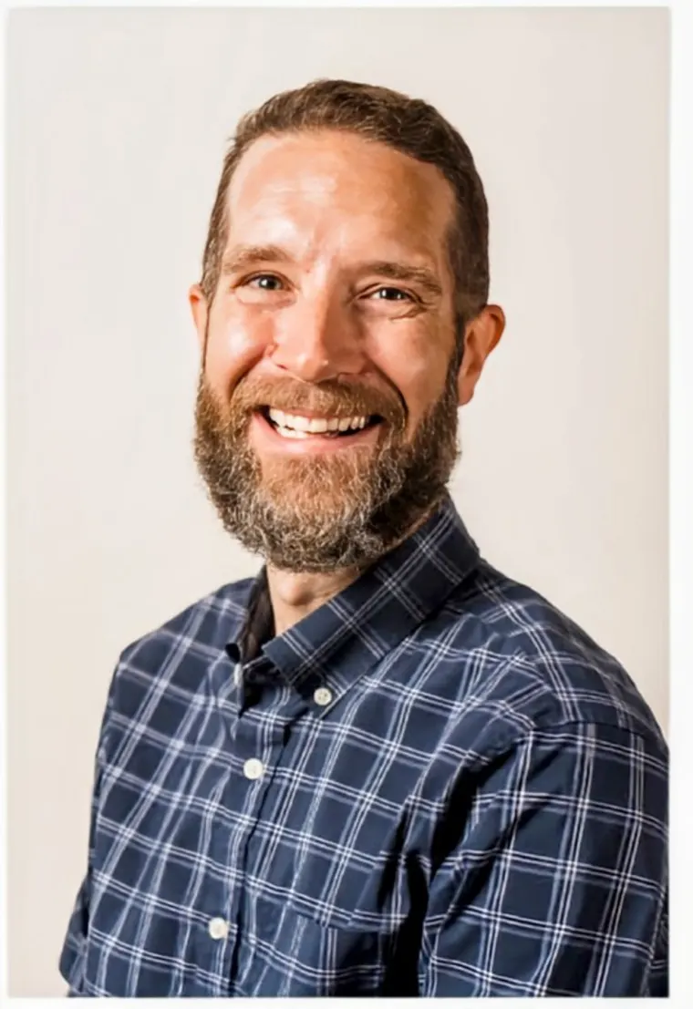 Person smiling with a beard, wearing a blue checkered shirt, posing against a plain background.