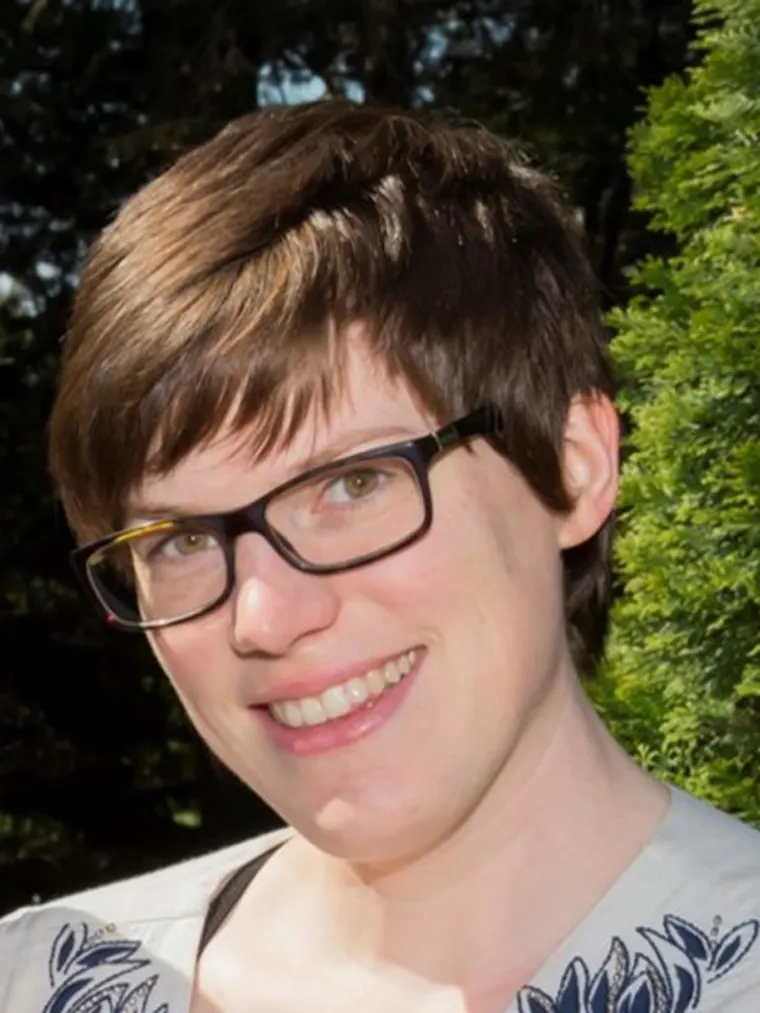 A person with short hair and glasses smiles outdoors, standing near green foliage under bright sunlight.