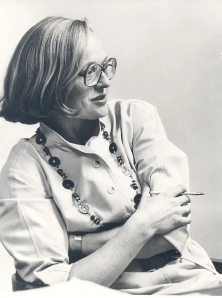Person wearing glasses and a beaded necklace, sitting with crossed arms and holding a pen, looking to the side. Black and white photo.