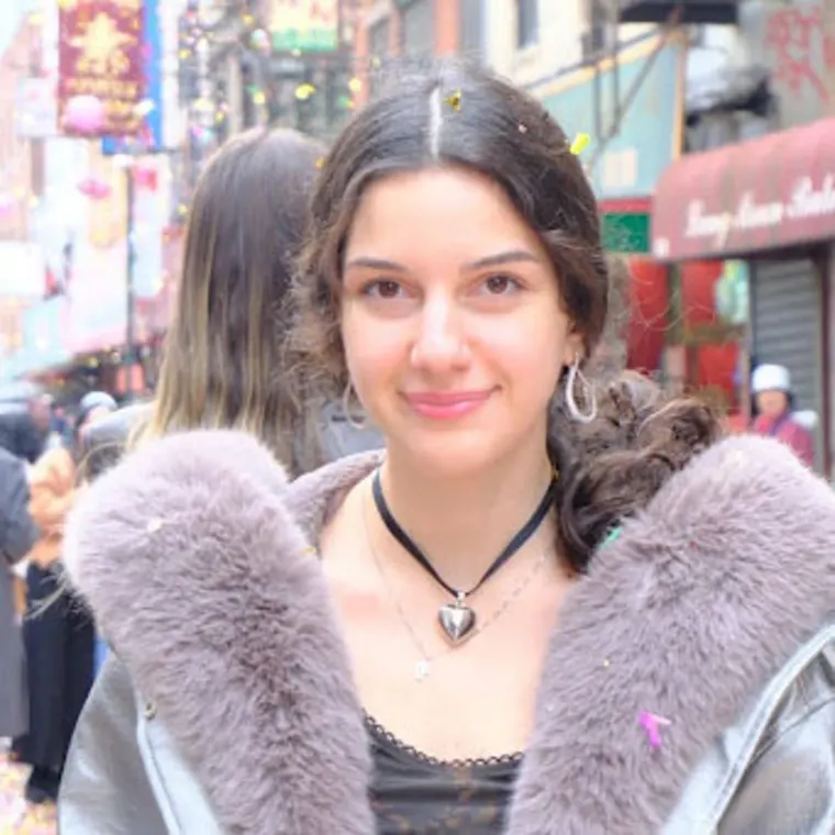 Person smiling, wearing a fur-collared coat and heart necklace, stands on a busy street with colorful signs and lights in the background.