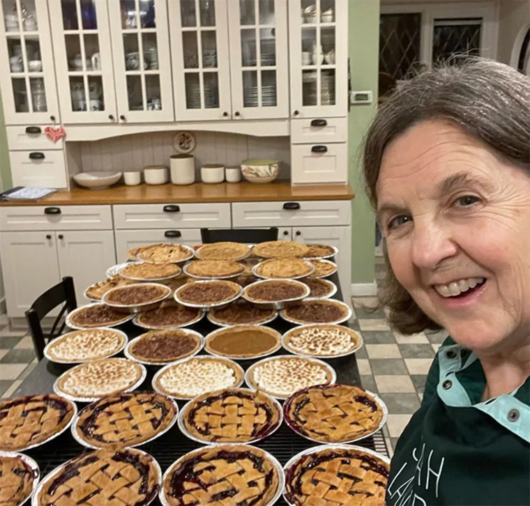 A person smiles next to a table filled with various pies. A kitchen with white cabinets is visible in the background.