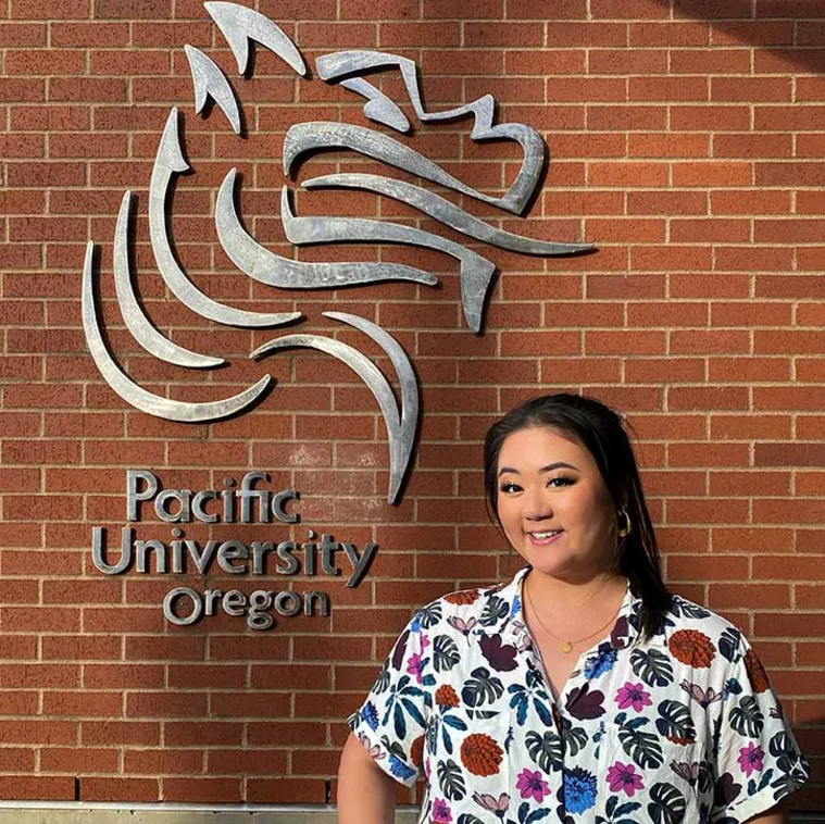 Person standing in front of a brick wall with the Pacific University Oregon logo, wearing a floral patterned shirt, smiling.