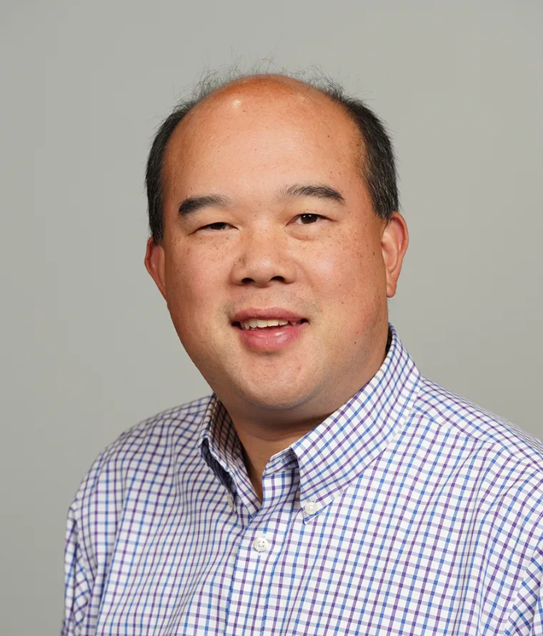 Person smiling, wearing a checked shirt, with a neutral background.