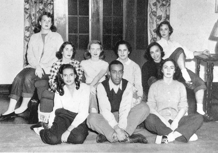 Nine people posing in a vintage group photo, sitting and standing in a living room with a window in the background.