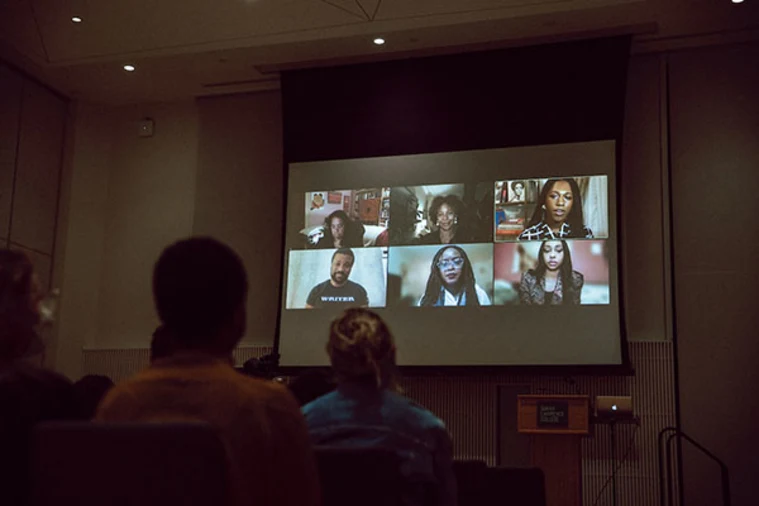 Audience watching a video conference on a large screen showing six participants in a dimly lit room.