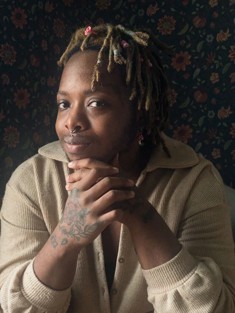Person with dreadlocks and piercings, resting chin on hands with tattooed floral wallpaper background.