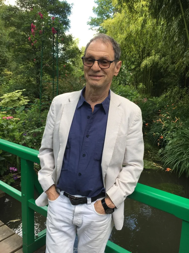 Person wearing glasses, white jacket, and dark shirt, standing on a bridge against a lush green garden backdrop.