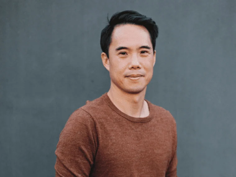 Person in a brown sweater stands in front of a gray background with a slight smile.