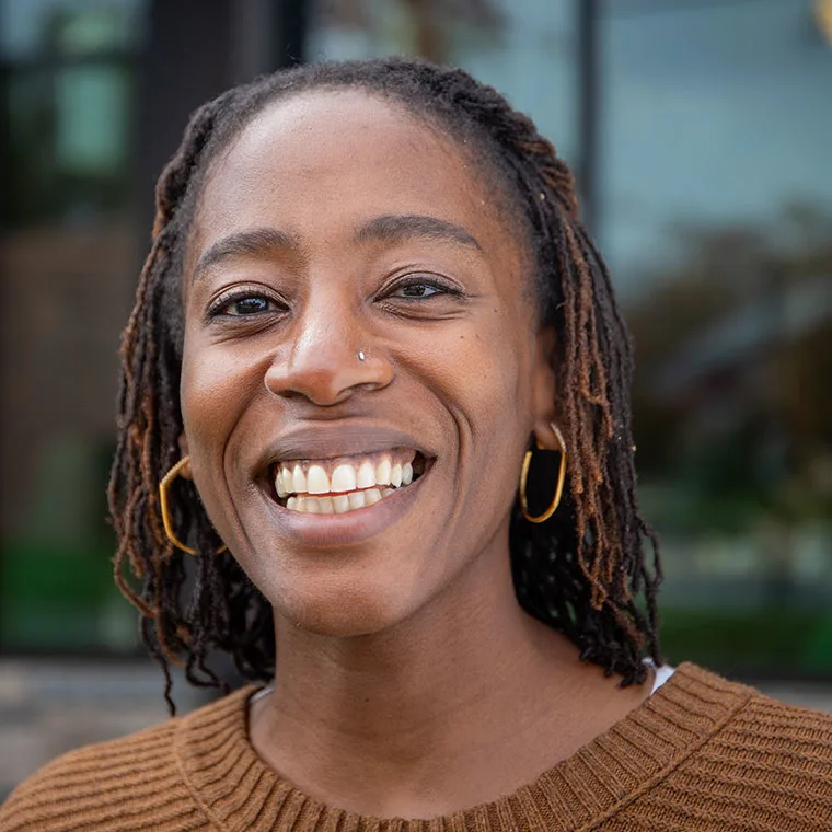 Person with braided hair, wearing hoop earrings and a brown sweater, smiling in front of a blurred outdoor background.