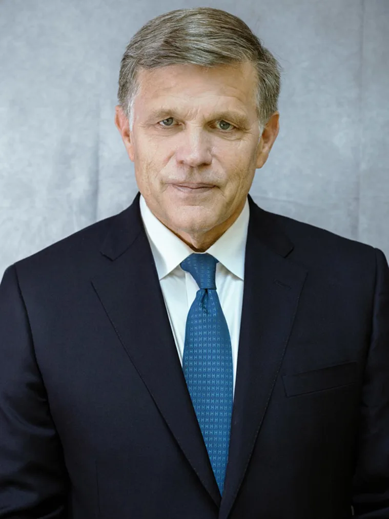 Person in a dark suit and blue tie, with short hair, against a gray background.