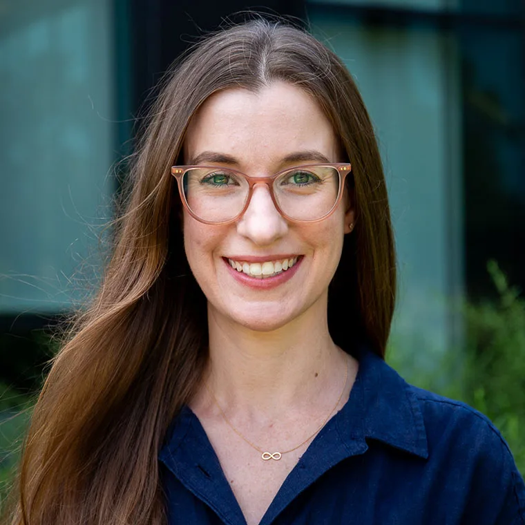 Person with long hair and glasses, smiling, wearing a blue shirt and infinity necklace, in front of a blurred outdoor background.