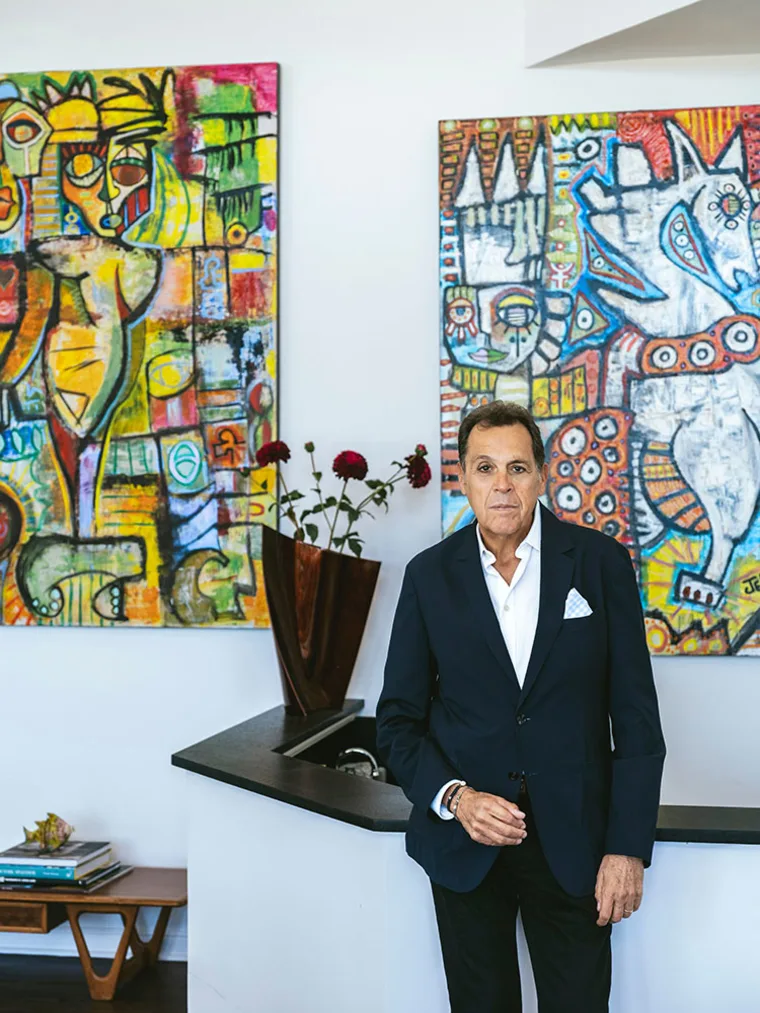 Person in a suit stands near a counter with abstract artwork in the background and a vase with red flowers.