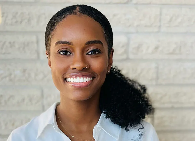 Person smiling in front of a light brick wall, wearing a white shirt and a nose ring, with curly hair pulled back.