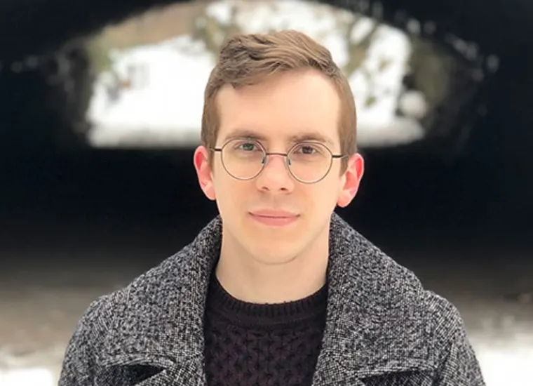Person with glasses in a thick coat stands outdoors, with a snowy background visible through a tunnel.